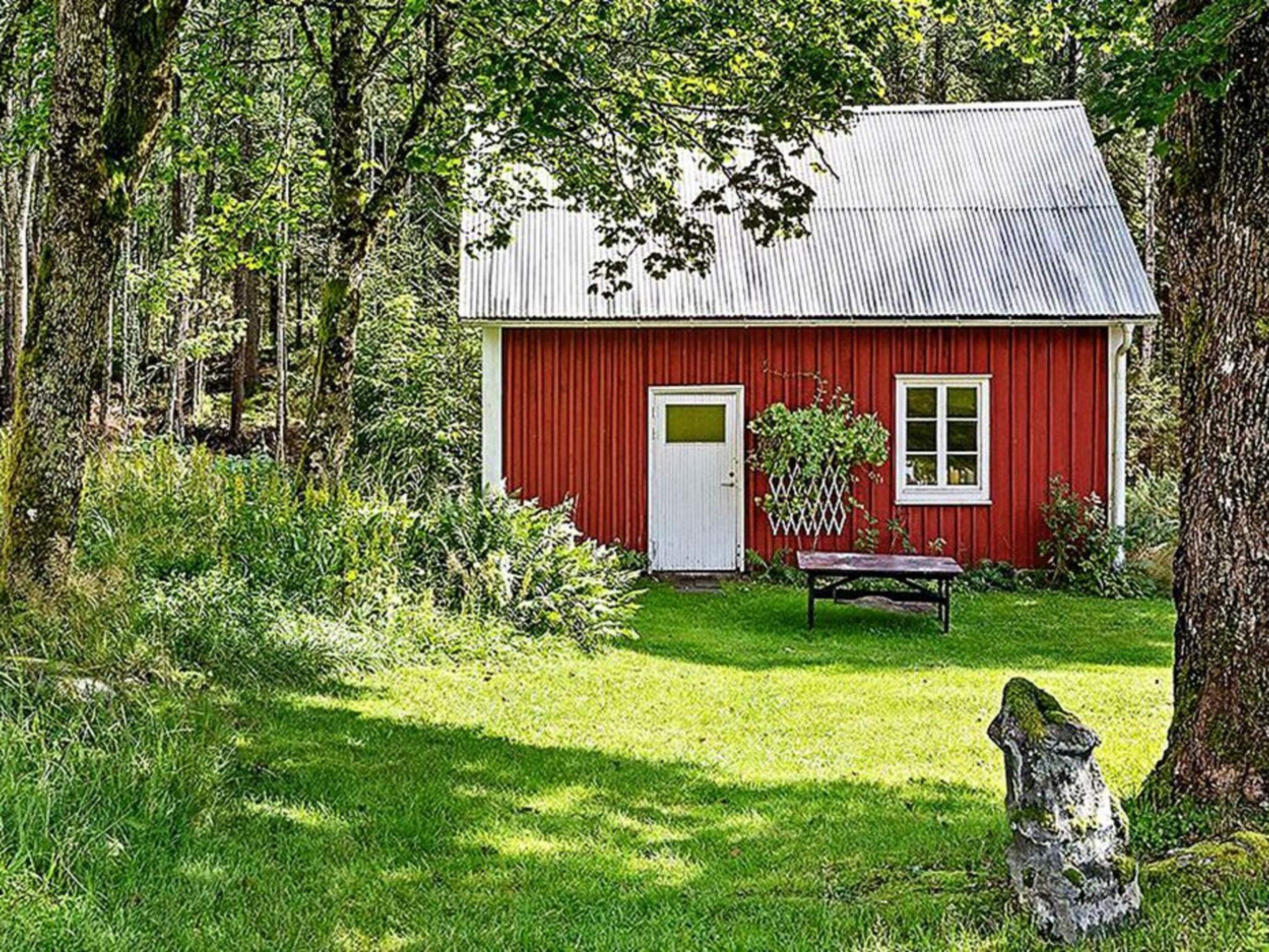 Une maison en bois rouge, avec une porte et une fenêtre blanches, entourée d’une cour verdoyante et d’arbres.
