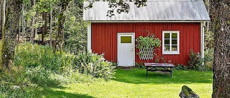 A red wooden house with a white door and window, surrounded by a grassy yard and trees.