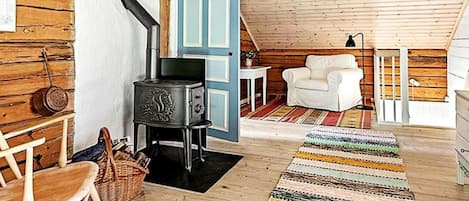 A cozy attic room with a wooden ceiling, a black stove, a white rocking chair, a white chair, and a colorful striped rug.
