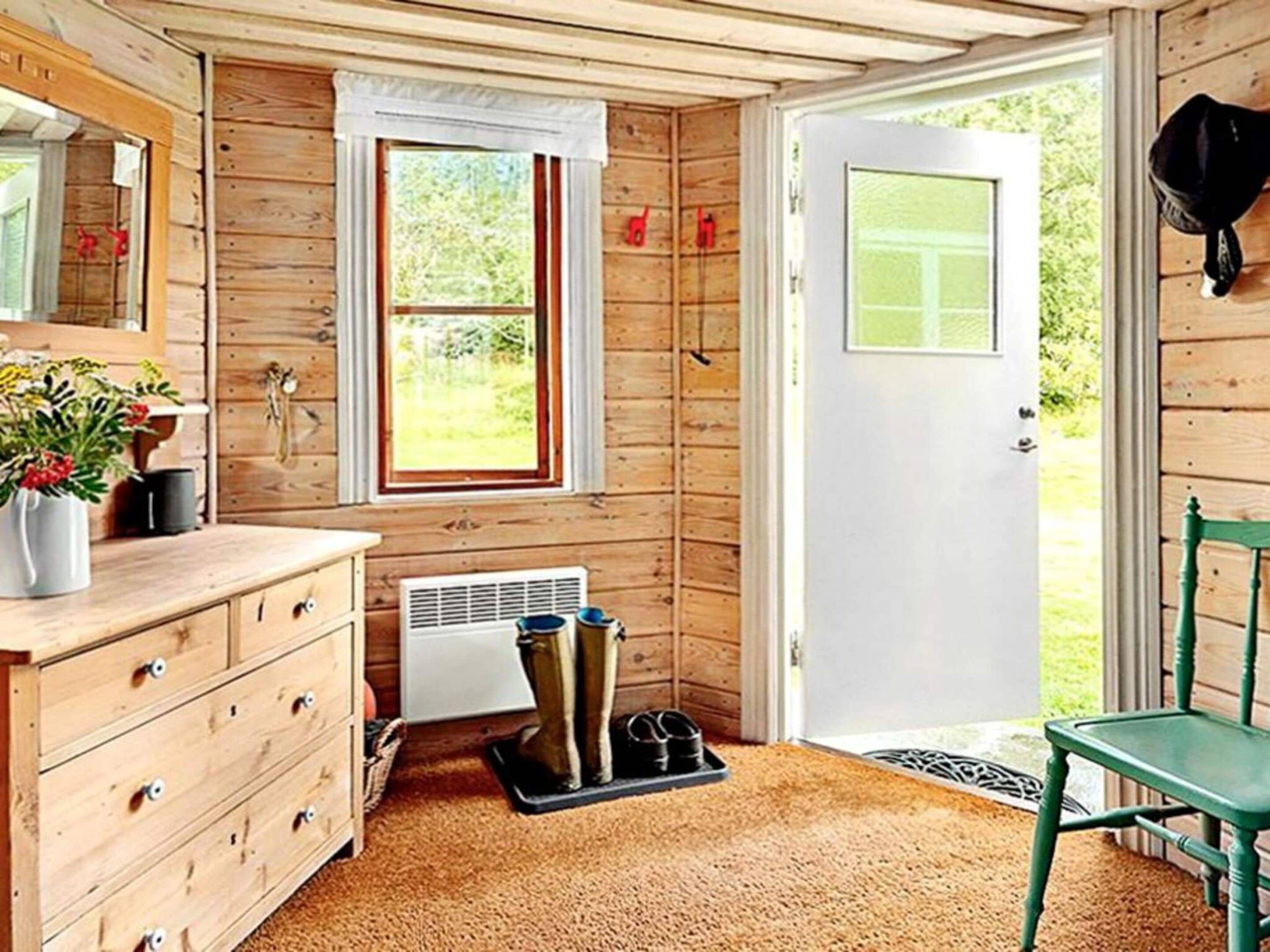 A wooden interior with a wooden dresser, a window with curtains, a radiator, a door, and a green chair.