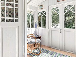 A room with a white door, a wicker chair, a small table, and a window with a view of trees. - 4 Star Holiday Home in Skillingaryd (Skillingaryd)