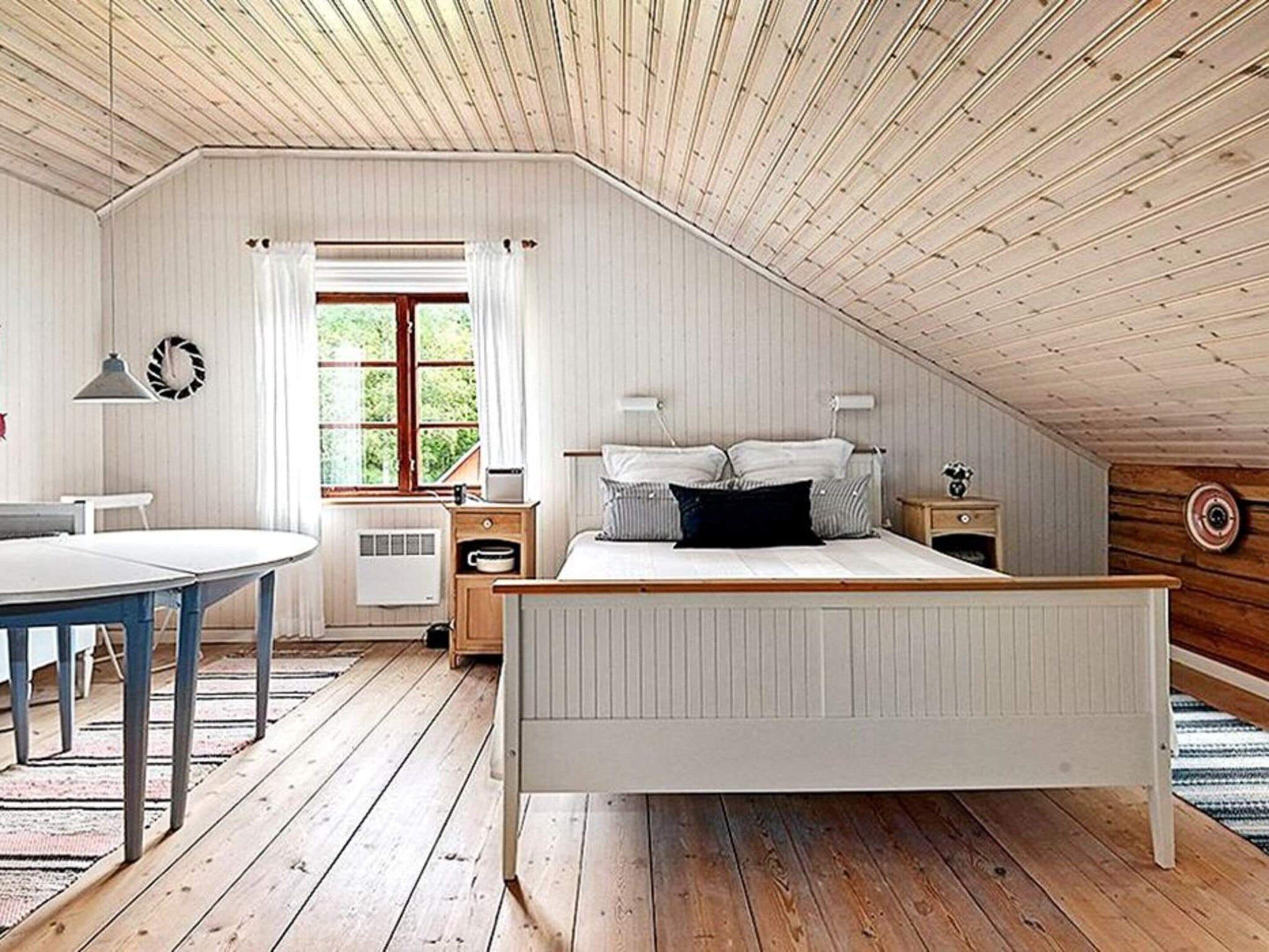 A bedroom with a sloped ceiling, a bed with white bedding, a bedside table, a lamp, and a window with curtains.