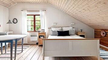 A bedroom with a sloped ceiling, a bed with white bedding, a bedside table, a lamp, and a window with curtains.
