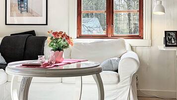 Una habitación con un sofá blanco, una mesa redonda con un jarrón de flores, una ventana con vistas a árboles y un cuadro en la pared.