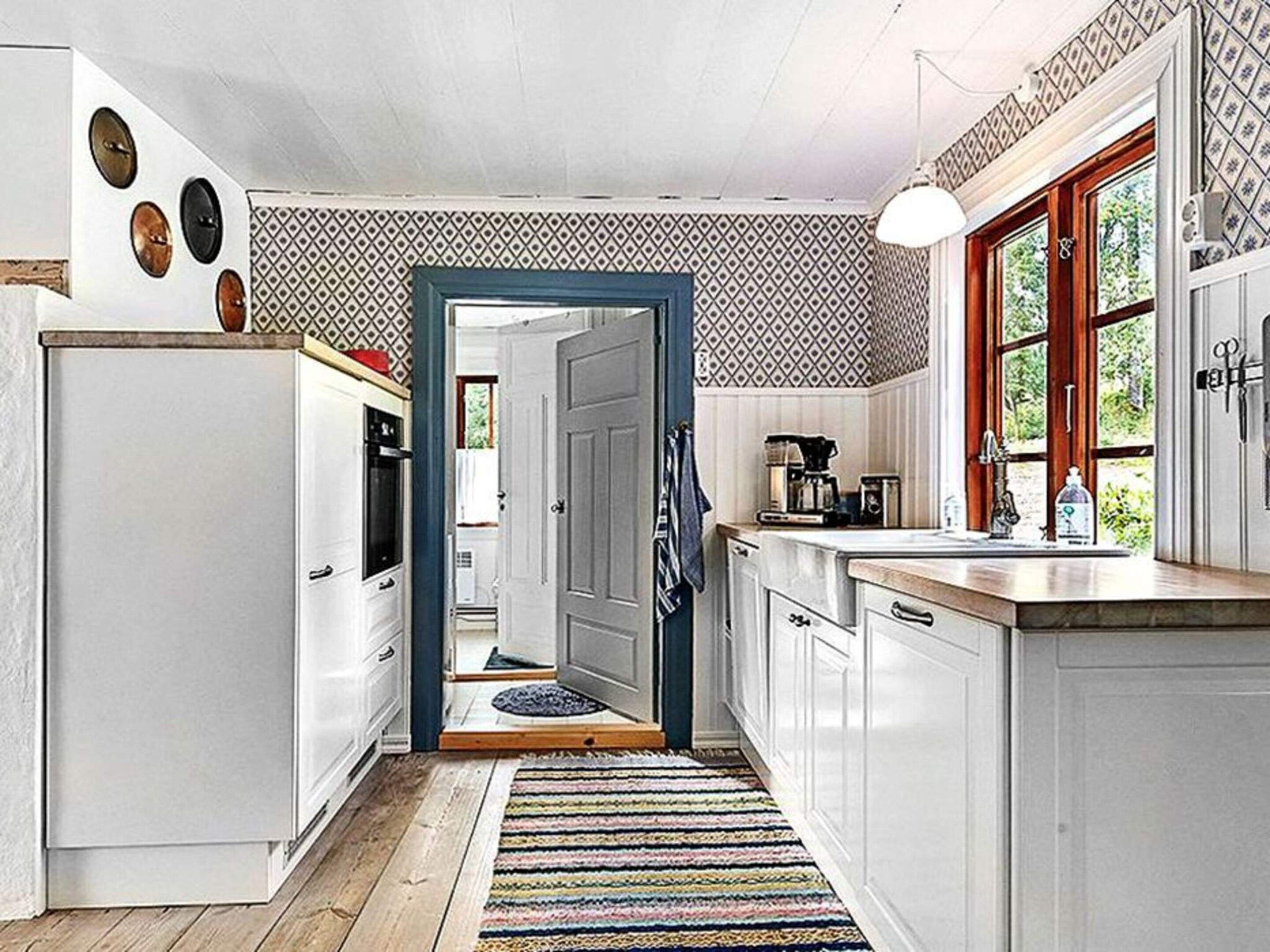 Una cocina con armarios blancos, piso de madera, alfombra colorida y ventana con vistas a zonas verdes.