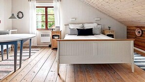 Ein Schlafzimmer mit schrägen Wänden, einem Bett mit weißer Bettwäsche, einem Nachttisch, einer Lampe und einem Fenster mit Vorhängen.