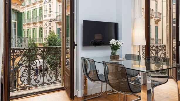 Un espace repas moderne avec une table et des chaises en verre, un téléviseur à écran plat fixé au mur et un balcon agrémenté de balustrades en fer forgé.