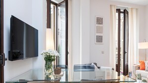 Un salon moderne avec une table à manger en verre, un téléviseur à écran plat fixé au mur et un canapé agrémenté de coussins.