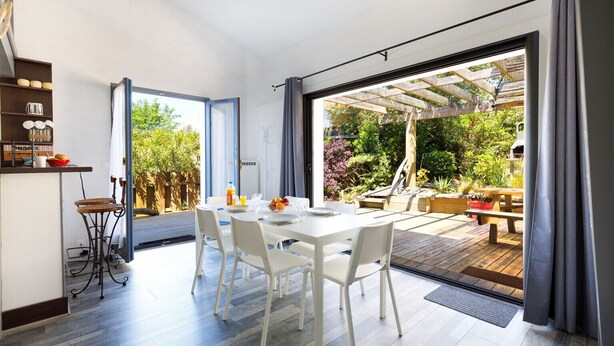 Una cocina moderna con armarios blancos, un comedor con mesa y sillas blancas, y un patio con terraza de madera y mobiliario exterior.