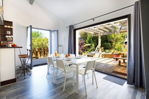 Una cocina moderna con armarios blancos, un comedor con mesa y sillas blancas, y un patio con terraza de madera y mobiliario exterior.