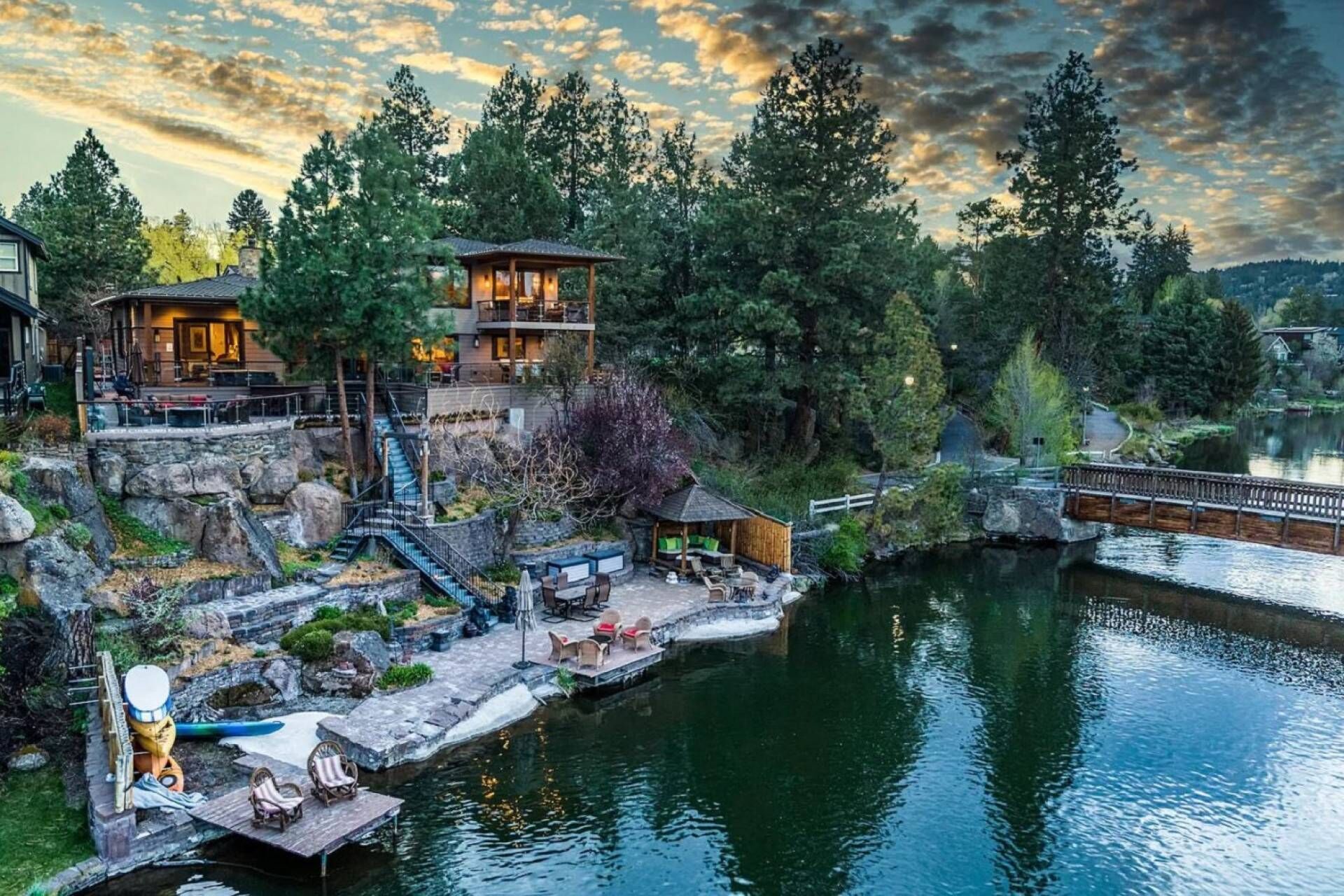 A lakeside lodge with a stone staircase leading to a wooden deck, a dock with chairs, and a bridge over the water.