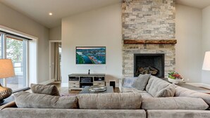 Una sala de estar con chimenea de piedra, un televisor de pantalla plana grande montado encima, un sofá seccional y un mueble de entretenimiento de madera con dispositivos electrónicos.