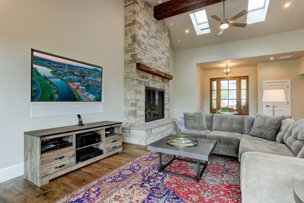 A living room with a stone fireplace, a gray sectional sofa, a colorful rug, and a mounted TV.