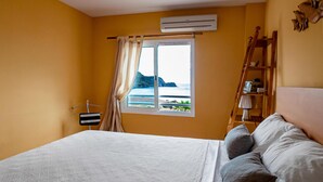 A bedroom with a bed, a window with curtains, an air conditioning unit, a wooden ladder, and a view of the sea. - Casa La Isleta     . (Níjar)