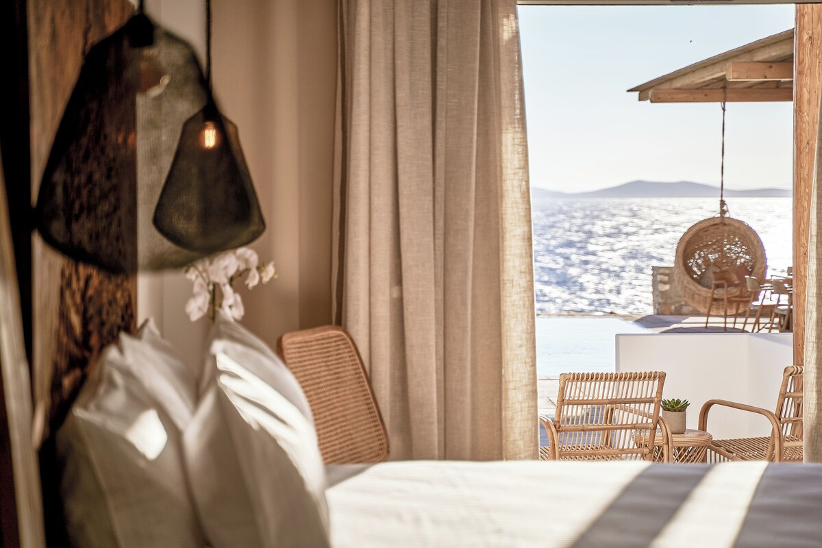 A bedroom with a bed, a hanging lamp, a chair, and a view of the sea.