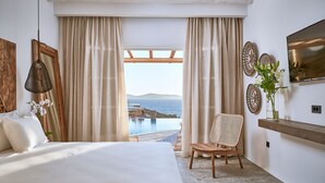A bedroom with a large bed, a chair, a view of the sea, and a mounted TV. - Ubud Mykonos (Mykonos)