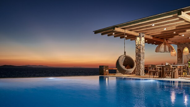 A poolside area with a view of the ocean at sunset, wooden pergola, stone pillars, and hanging wicker chairs. - Ubud Mykonos (Mykonos)