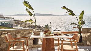 A wooden table set with plates, bowls, and a bottle, overlooking a scenic coastal view. - Ubud Mykonos (Mykonos)
