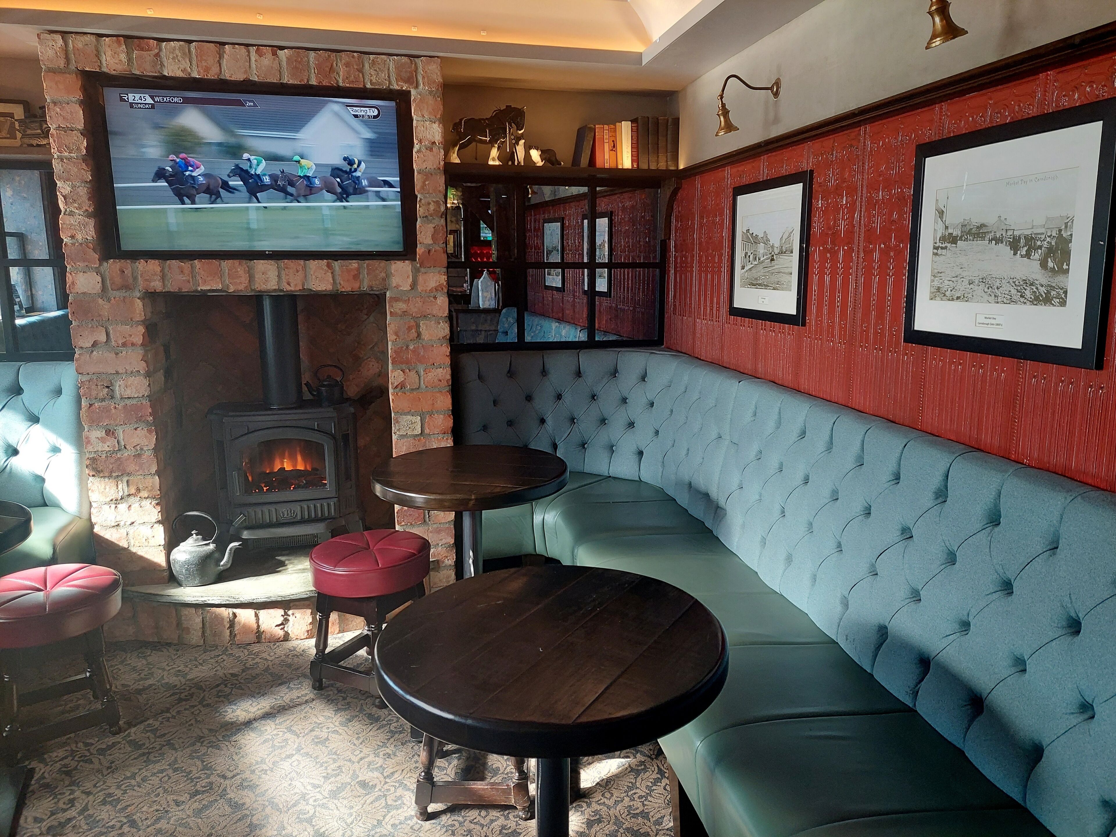 Un rincón de bar acogedor con chimenea, un televisor mostrando una carrera de caballos y una pared roja decorada con cuadros.