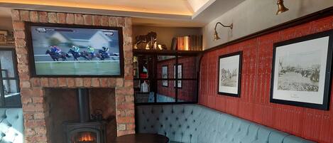 Un rincón de bar acogedor con chimenea, un televisor mostrando una carrera de caballos y una pared roja decorada con cuadros.