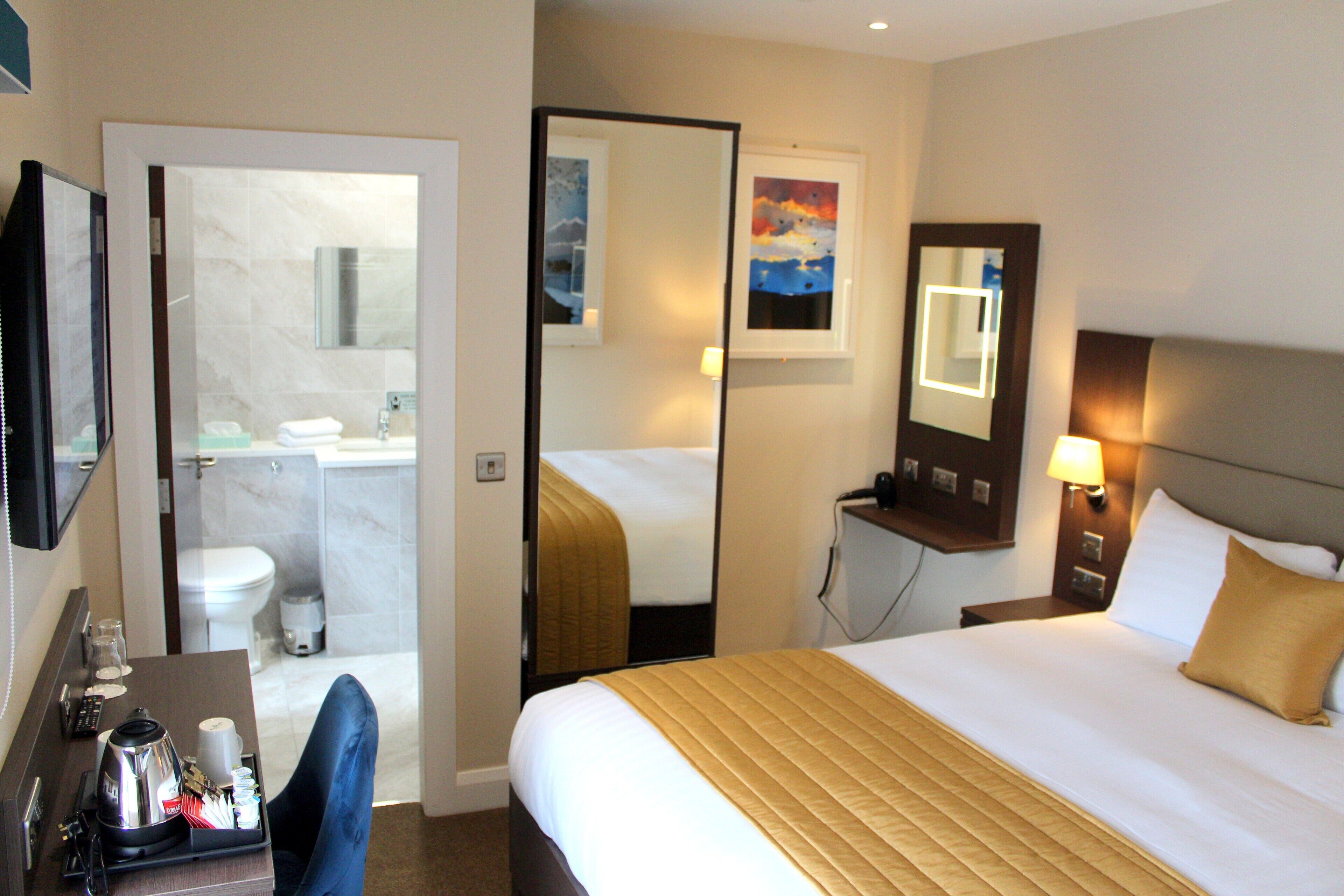 A hotel room with a bed, a desk with a coffee maker, a mirror, and a bathroom visible through an open door.