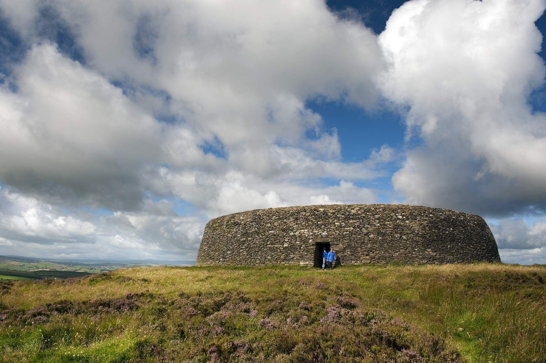 A stone structure with a circular entrance, surrounded by grassy terrain and under a partly cloudy sky.