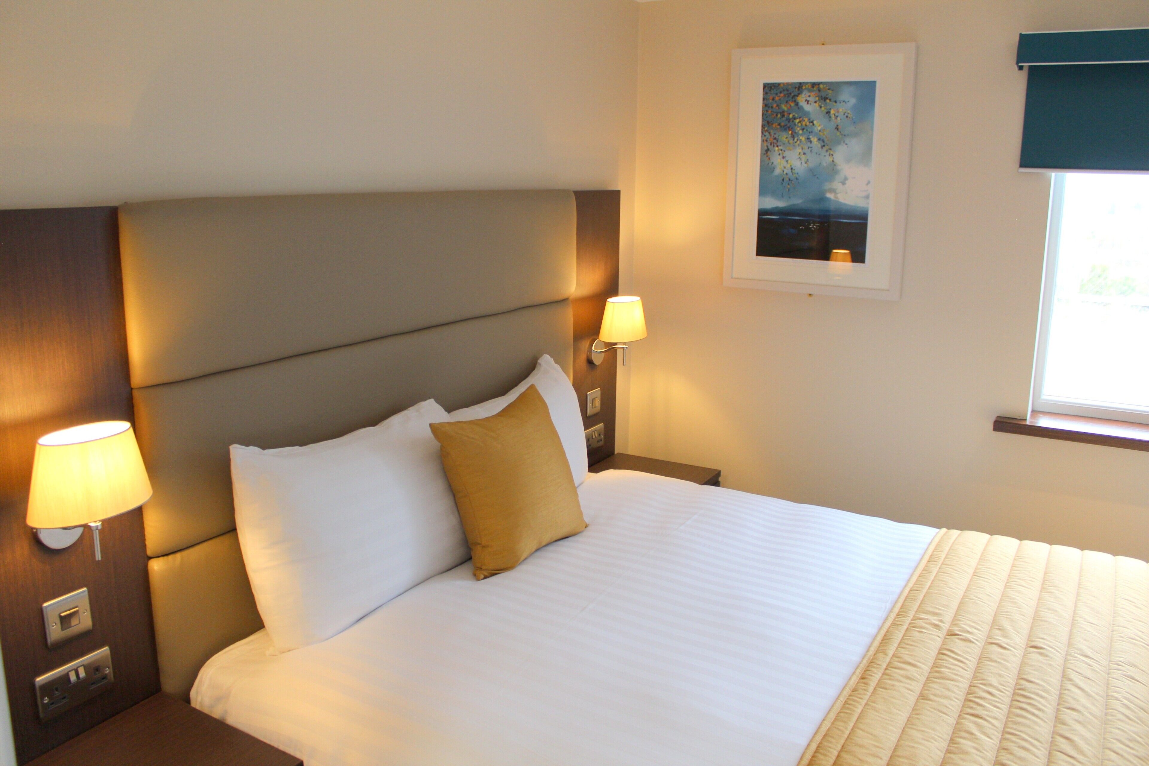 A neatly made bed with white and yellow pillows, a headboard, bedside lamps, and a framed picture on the wall.