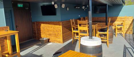 An outdoor seating area with wooden tables and chairs, a large barrel, and a door leading to the interior.