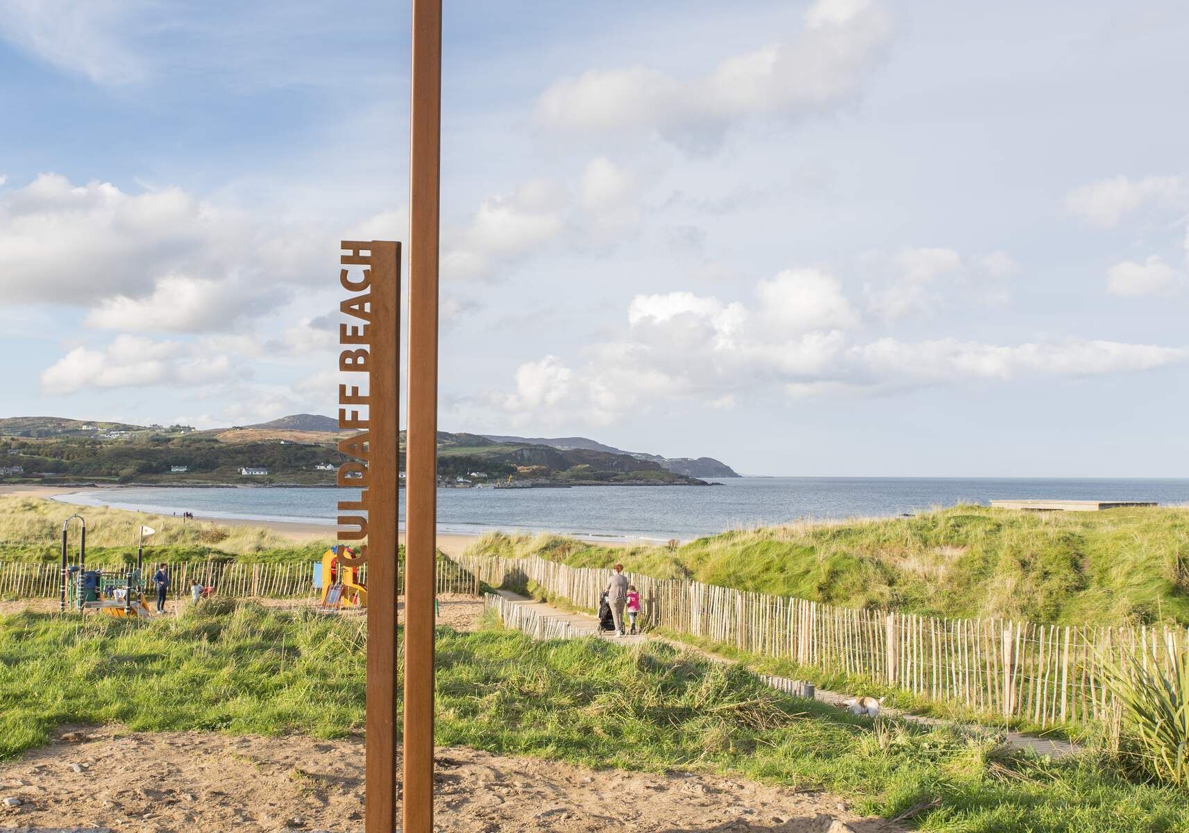 Un cartello di legno con su scritto "Culdee Beach", una spiaggia sabbiosa con una recinzione in legno e una zona verde con persone e un cane.