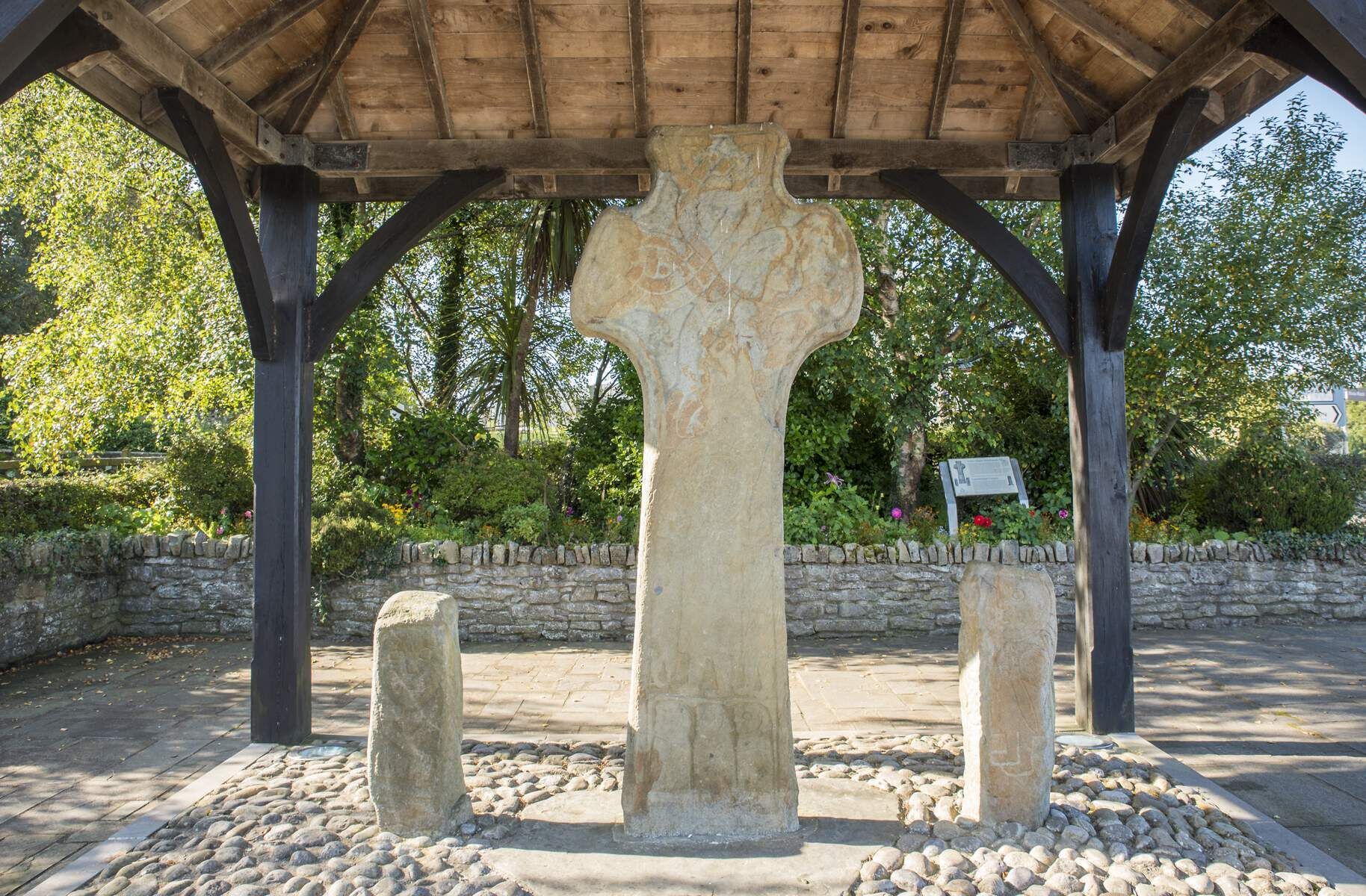 Une croix en pierre, placée sous un pavillon en bois soutenu par des piliers de pierre, est entourée d’un mur de pierre et de verdure.