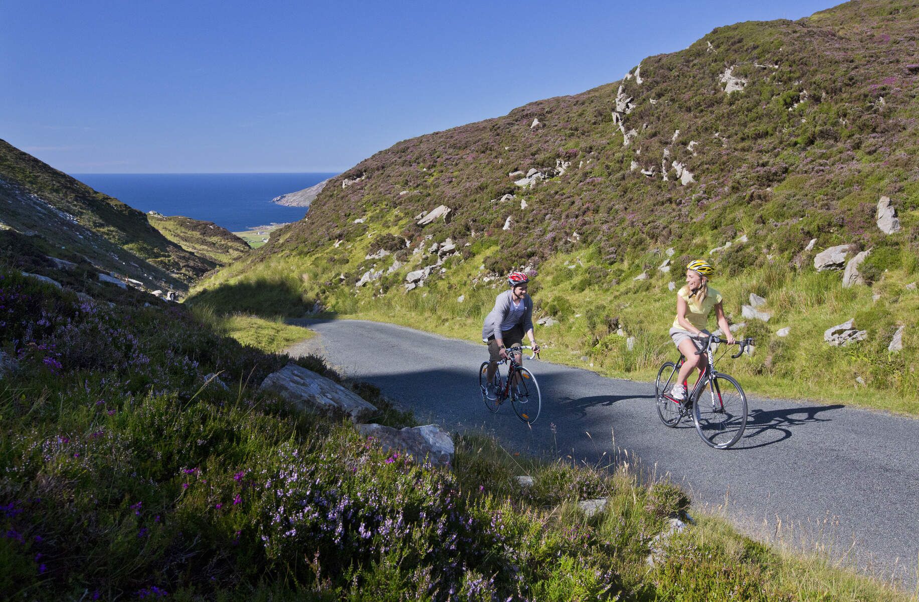 Two cyclists on a narrow road surrounded by hills and wildflowers.