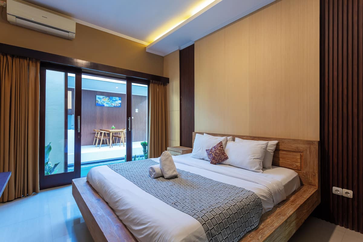 A hotel room with a large bed, a wooden headboard, a sliding glass door leading to a balcony, and a small table with chairs outside.