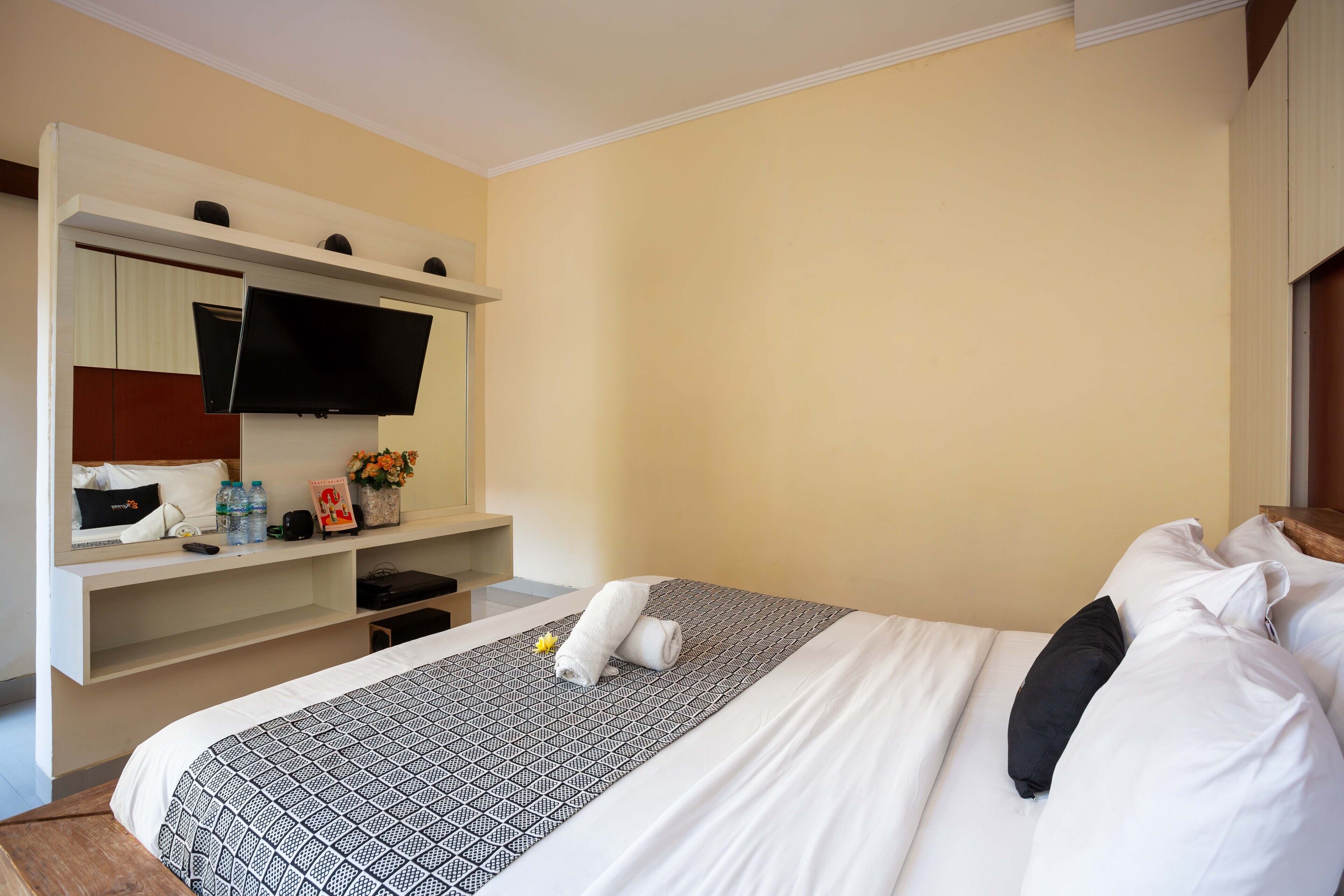 A hotel room with a bed, a TV mounted on a wall, a desk with a chair, and a shelf with toiletries.