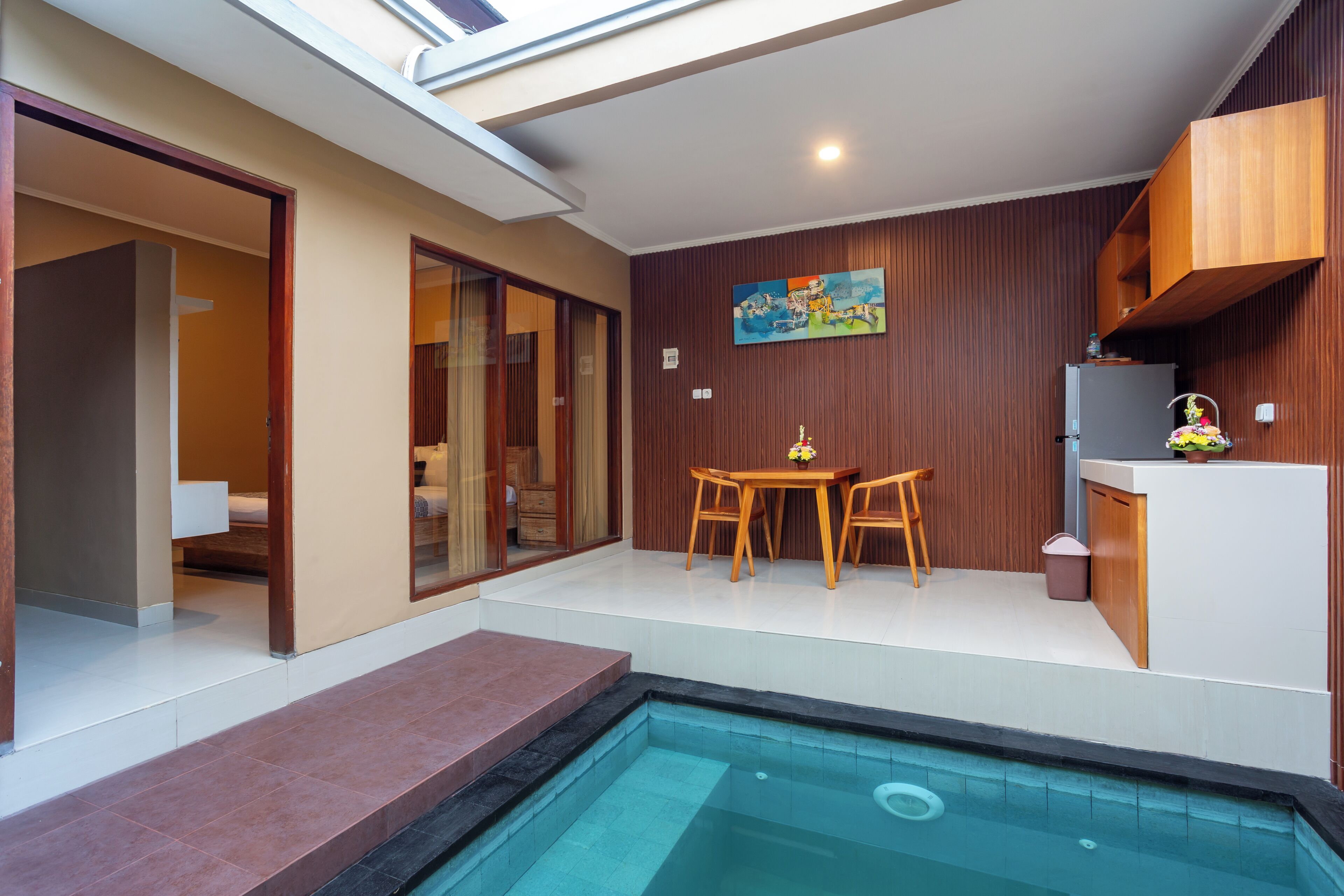 A modern indoor pool area with a dining table and chairs, a small kitchen area, and a painting on the wall.