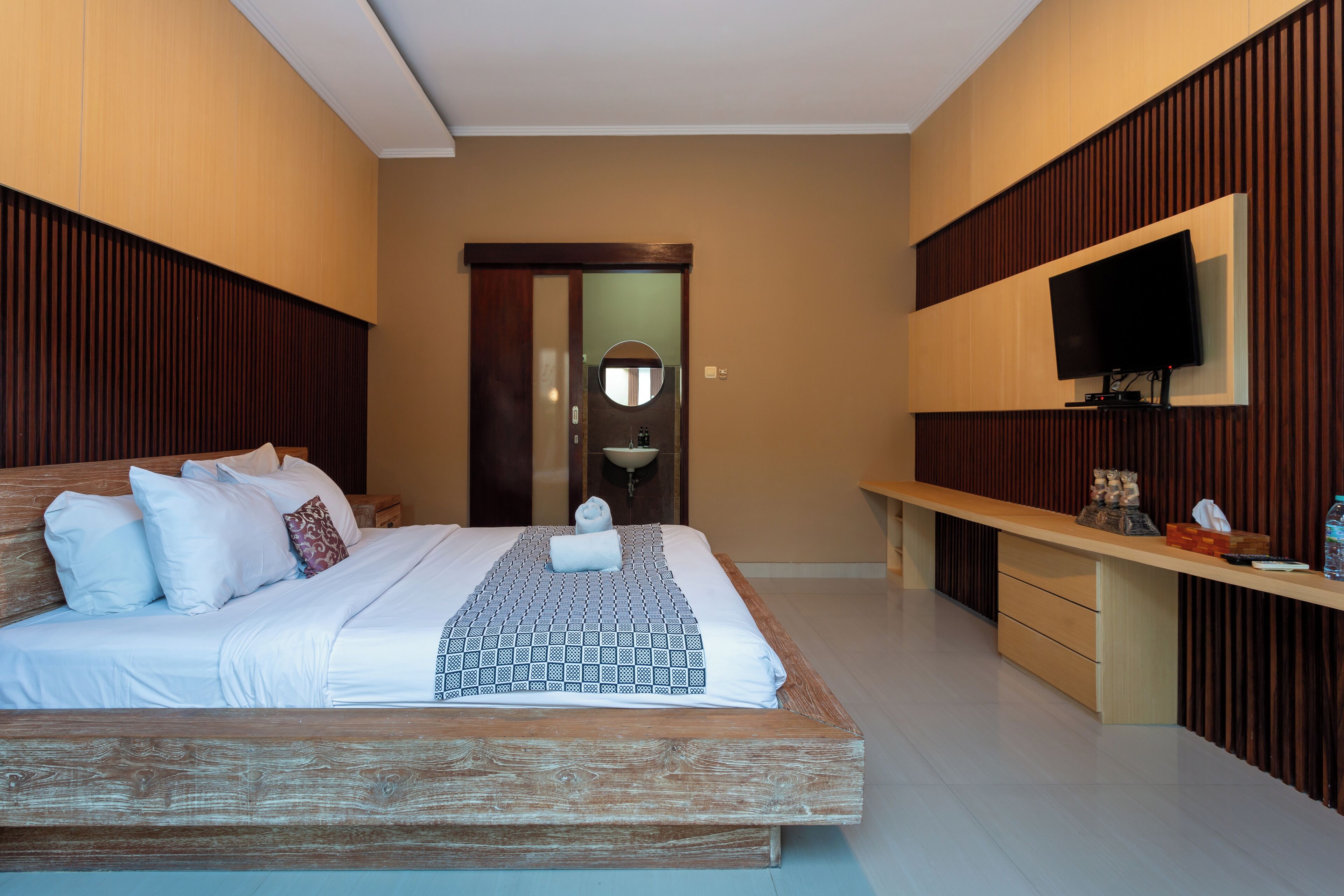 A modern hotel room with a wooden bed, a flat-screen TV mounted on the wall, and a small desk with a mirror and a bottle of water.
