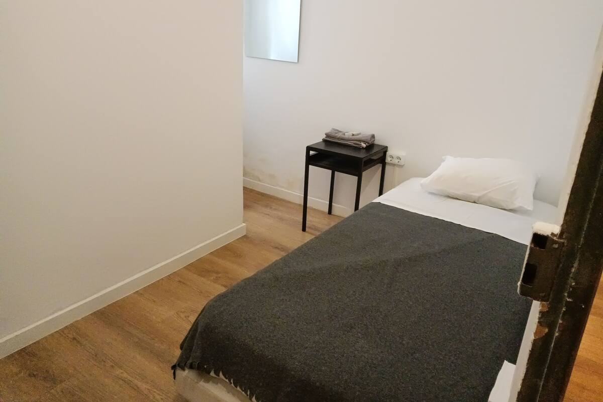 Ein kleines, schlichtes Zimmer mit einem Bett, einem kleinen Tisch und einem Spiegel an der Wand.