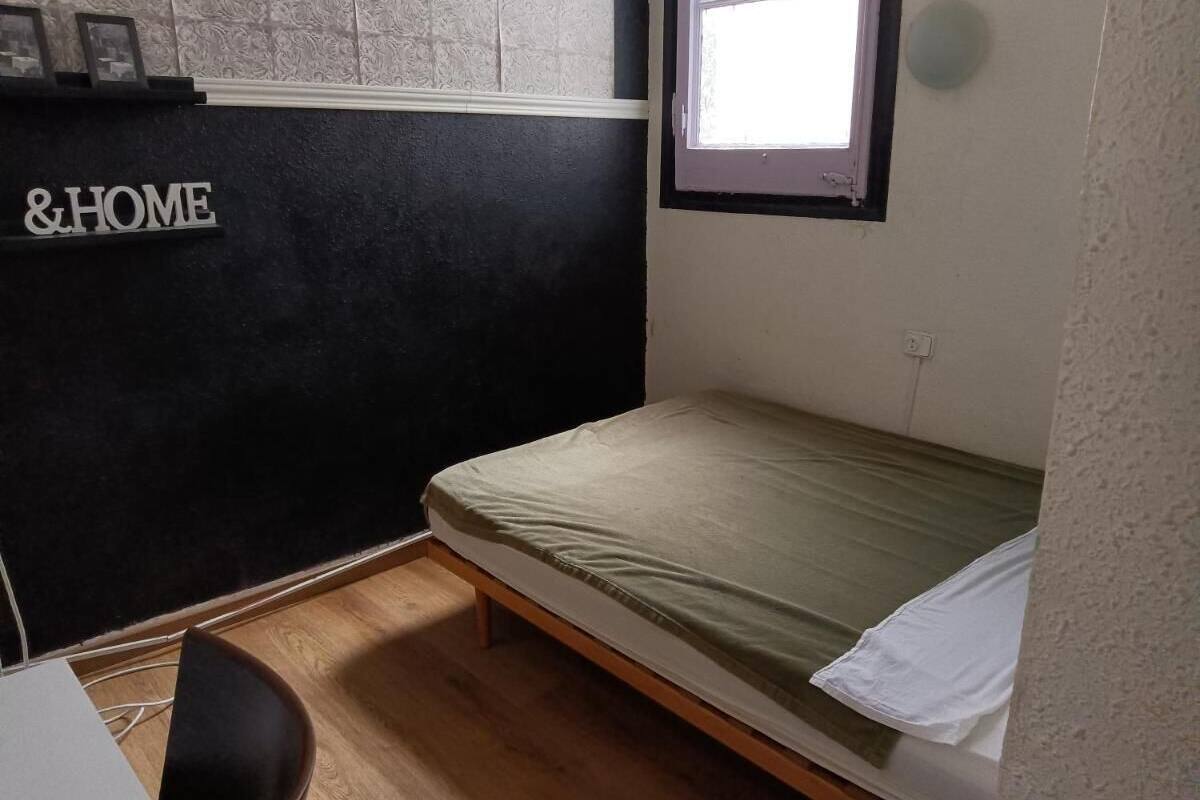 Ein kleines Zimmer mit einem Einzelbett, Holzboden, einem Fenster und einem an der Wand befestigten Regal mit einem Schild, auf dem '&HOME' steht.