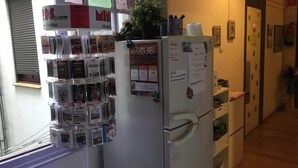 Una cocina con un refrigerador que muestra varios imanes y notas, una ventana con persianas y una puerta que da a otra habitación.