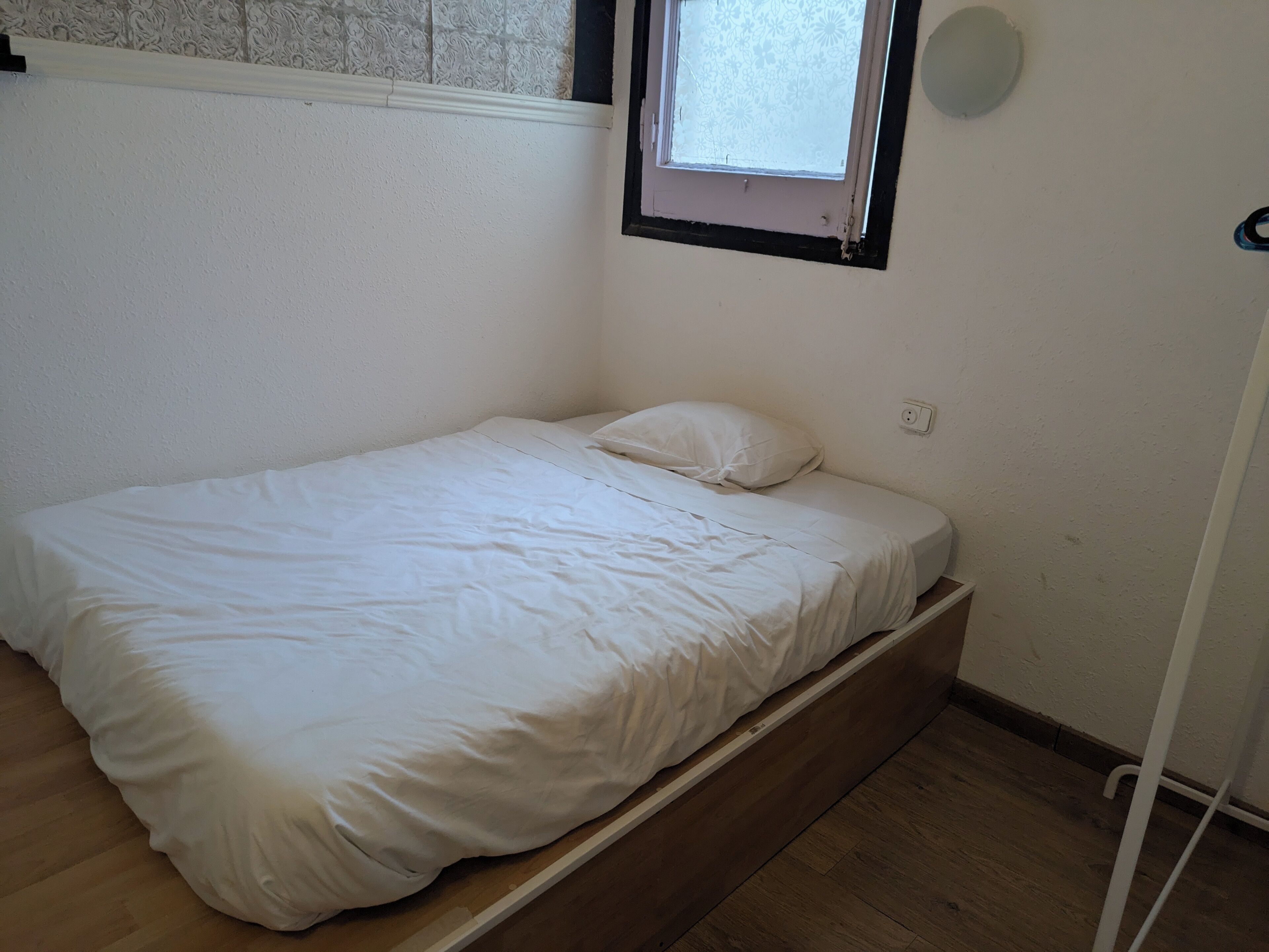 Una habitación pequeña con cama individual, suelo de madera, paredes blancas y una ventana con persiana estampada.