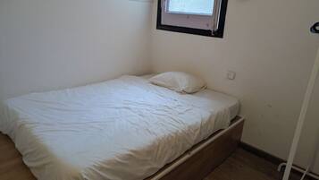 Una habitación pequeña con cama individual, suelo de madera, paredes blancas y una ventana con persiana estampada.