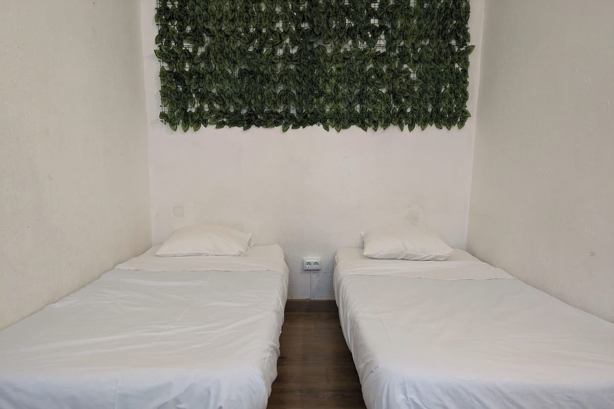 Ein Zimmer mit zwei Einzelbetten, weißer Bettwäsche, einem kleinen Nachttisch mit Lampe sowie einer Wanddekoration mit Grünpflanzen.