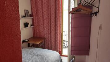 Ein kleines Schlafzimmer mit Bett, einem Holztisch, einer Kleiderstange und einem Fenster mit gemusterten Vorhängen.