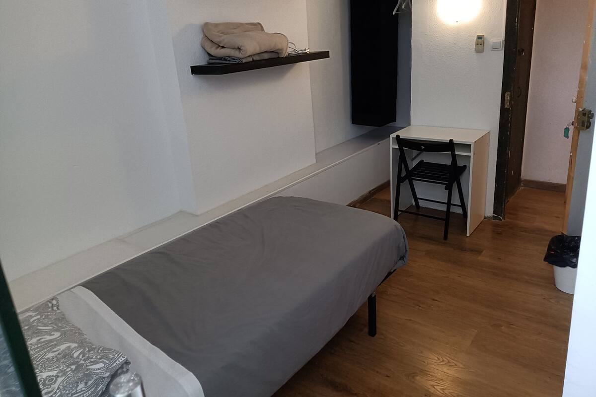 Ein kleines Zimmer mit einem Einzelbett, einem Schreibtisch und Stuhl, einem Regal mit einem Handtuch und einem an der Wand befestigten Fernseher.