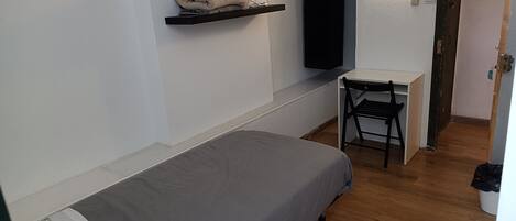 A small, single-bed room with a desk and chair, a shelf with a towel, and a wall-mounted TV.