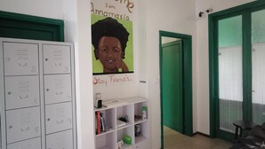 A room with a white wall, a green door, and a black table. There is a white cabinet with numbered compartments and a shelf with books and a green container. - Florence Hostels Anamaria (Florence)