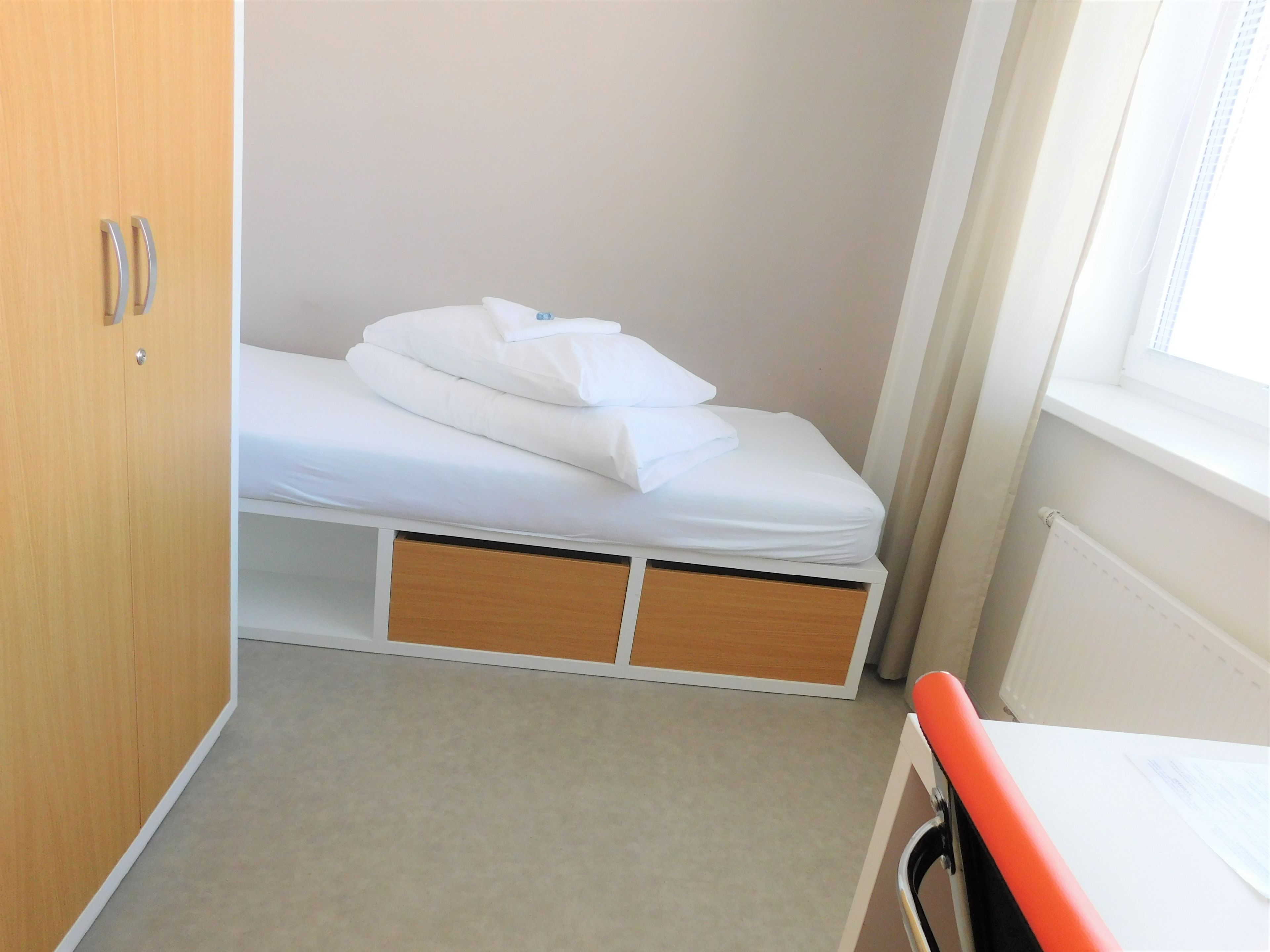 Una habitación pequeña con cama individual, un armario de madera, una ventana con persianas y un radiador.