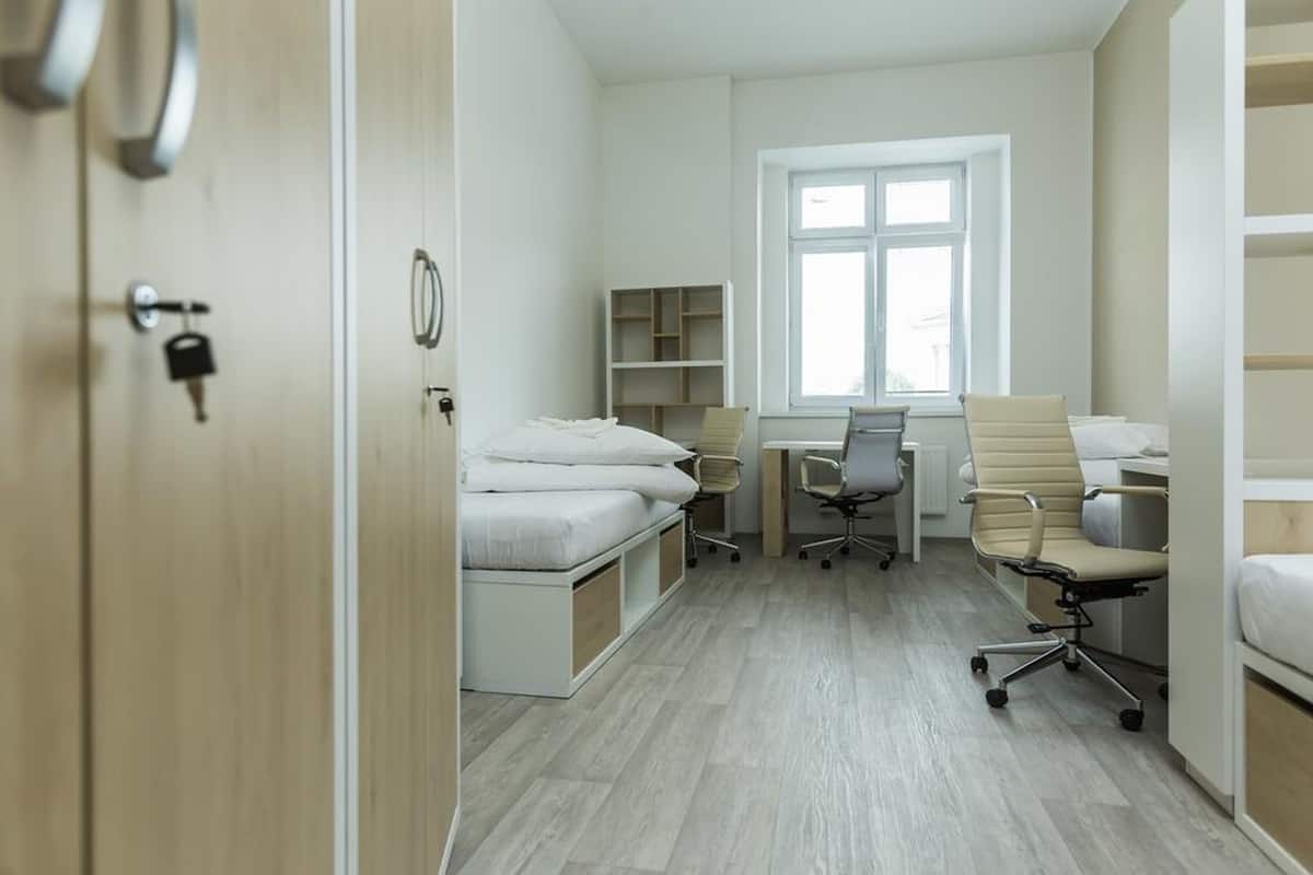 A compact room with a bed, desk, and chair, a window, and built-in shelving.