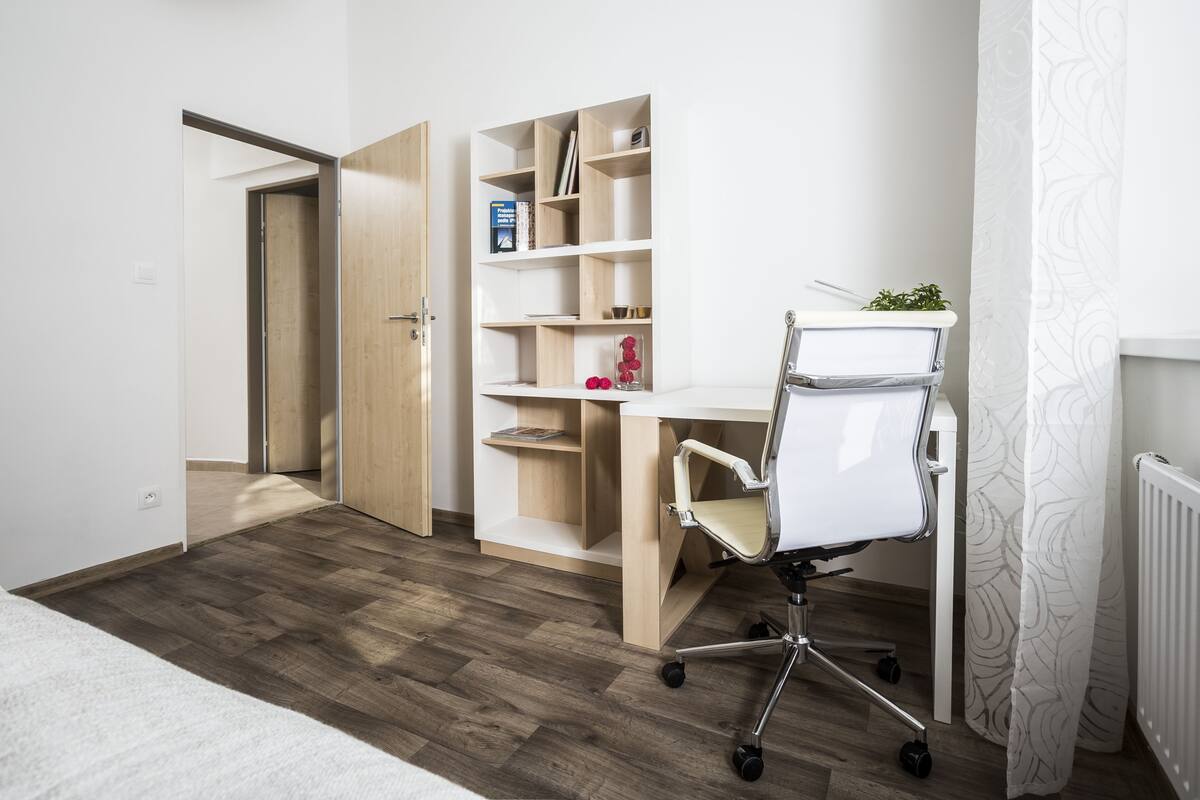 A modern bedroom with a wooden floor, a white bed, a desk with a chair, and a bookshelf.