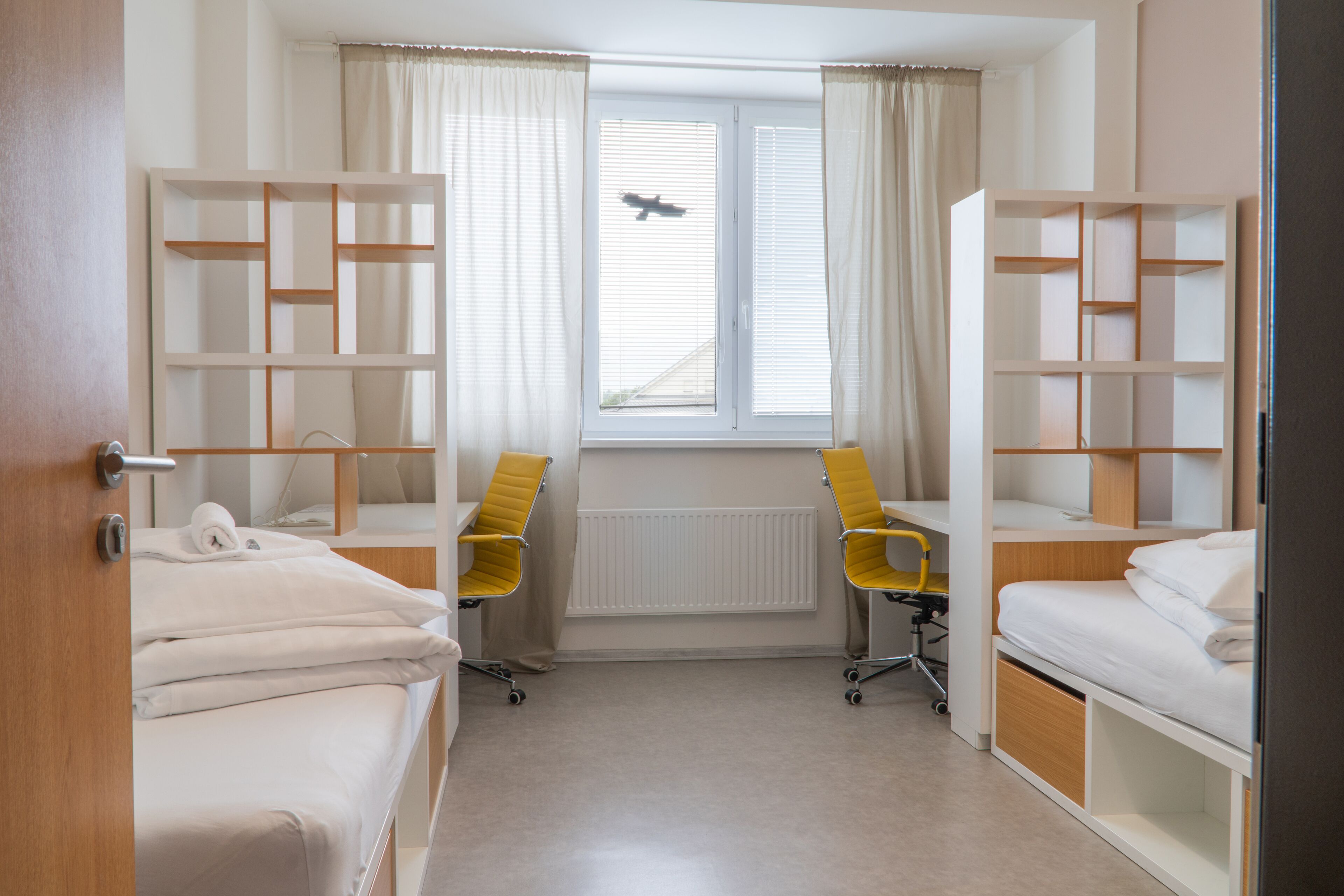 A hotel room with two single beds, a desk, two chairs, and a window with blinds.