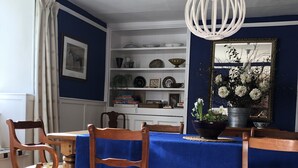 A dining area with a blue tablecloth, wooden chairs, a white chandelier, and a bookshelf with various decorative items.